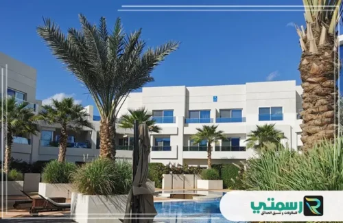 مبانٍ بيضاء حديثة تحيط بمسبح، مع نخيل كثيف وسماء زرقاء صافية.