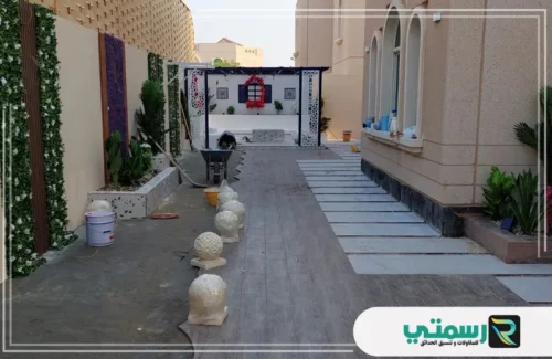 ساحة خارجية قيد التنسيق، بها مسار من البلاط، وكور حجرية، ومنطقة جلوس ببرجولا بيضاء.